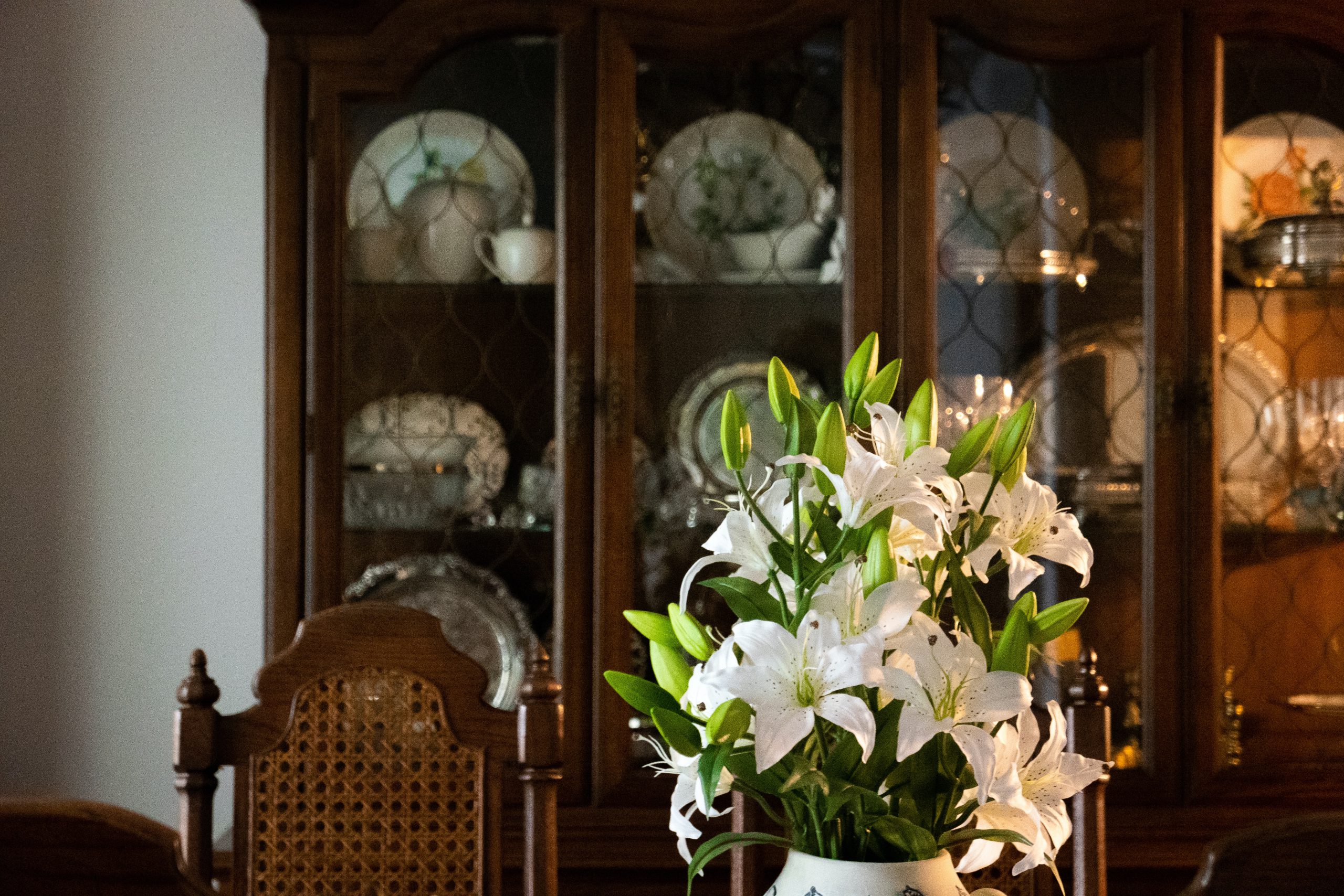 Dark wood china cabinet with a vase with white lilies in the foreground.