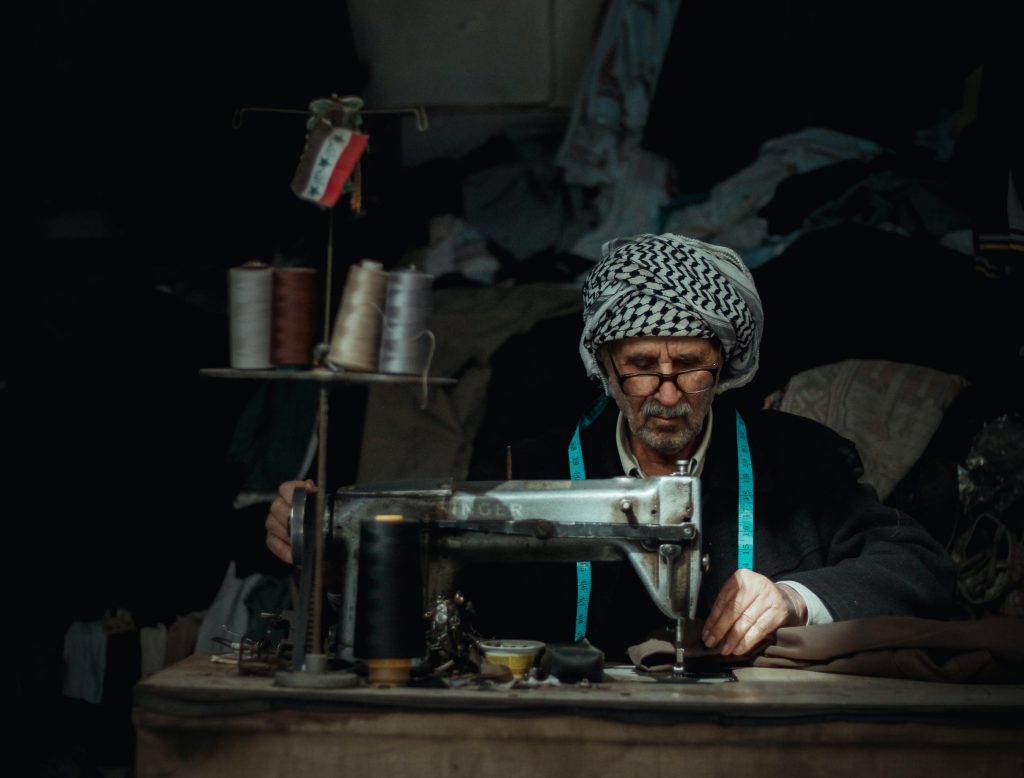 Old man wearing a black and white keffiyeh on his head sitting behind a singer sewing machine in a workshop