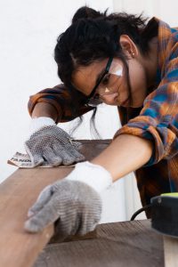 Woman wearing protective goggles and gloves sanding a wood plank